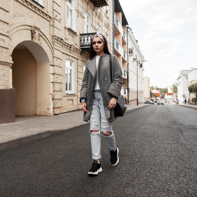 Moda giovane donna in bandana alla moda in cappotto elegante grigio in jeans strappati in scarpe da ginnastica con borsa nera a piedi vicino all'edificio d'epoca all'aperto. La ragazza adorabile in vestiti primaverili casuali della gioventù della moda gode del viaggio