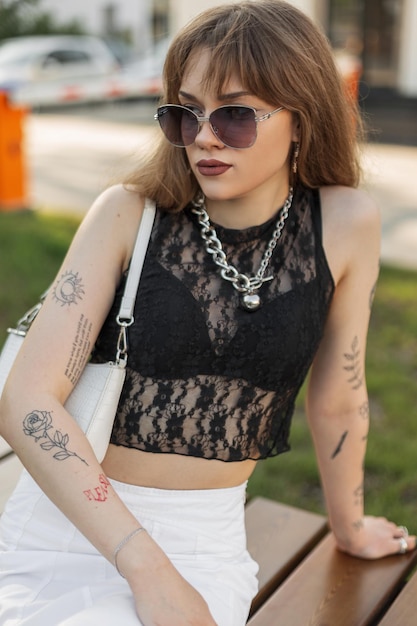 Moda hipster bellezza giovane donna con tatuaggi sulle braccia in abito estivo urbano alla moda con gonna in pizzo nero occhiali da sole e borsetta si siede sulla panchina