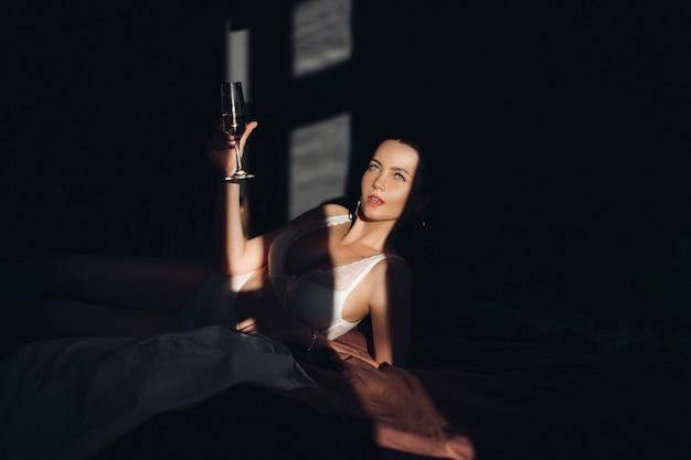 Moda ragazza bruna in lingerie sdraiata sul letto con bokal di champagne godendo di uno stile di vita di lusso
