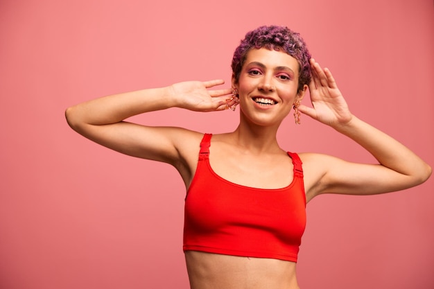 Moda ritratto di una donna con un taglio di capelli corto di colore viola e un sorriso con i denti in un top rosso su sfondo rosa felicità