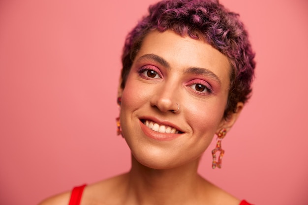 Moda ritratto di una donna con un taglio di capelli corto di colore viola e un sorriso con i denti in un top rosso su sfondo rosa felicità