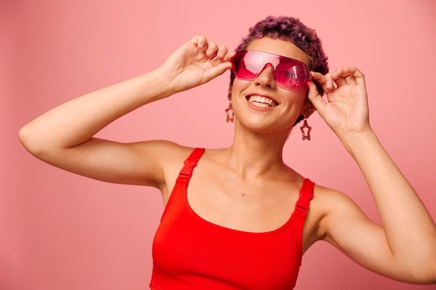 Moda ritratto di una donna con un taglio di capelli corto in occhiali da sole colorati e un sorriso con i denti in un top rosso su sfondo rosa