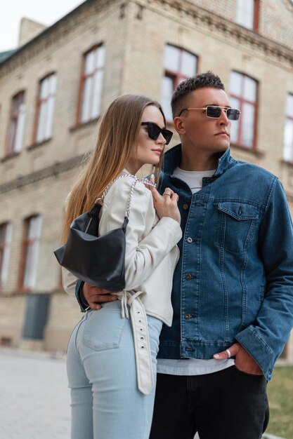 Moda street bella giovane coppia alla moda ragazzo e donna hipster con occhiali da sole vintage in abiti denim alla moda con una borsa che cammina insieme in città