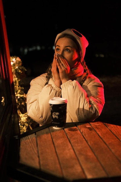 Modella bionda con la coda con una tazza di caffè vicino alla caffetteria con luce rossa e gialla cercando di scaldarsi le mani La ragazza indossava giacca e cappello bianchi in posa vicino al caffè nella notte d'inverno