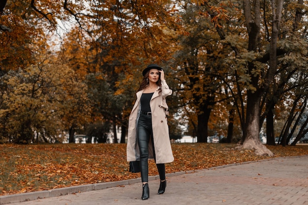 Modello alla moda bella ragazza con un cappello in un cappotto grigio alla moda con una borsa di moda passeggiate nel parco vicino agli alberi con fogliame autunnale giallo