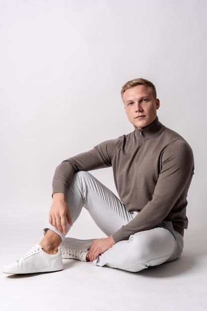 Modello biondo caucasico con un maglione marrone su sfondo bianco seduto sul pavimento con uno sguardo serio