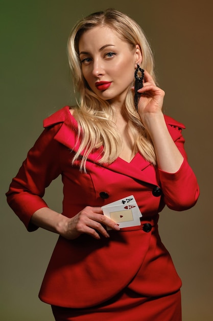 Modello biondo sveglio con trucco luminoso, in vestito rosso alla moda. Sta toccando il suo orecchino nero, mostrando due carte da gioco, in posa su sfondo colorato. Poker, casinò. Primo piano, copia spazio