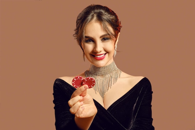 Modello bruna in abito nero e gioielli lucenti. Lei sorride, mostrando due fiches rosse, in posa su sfondo marrone. Poker, casinò. Avvicinamento