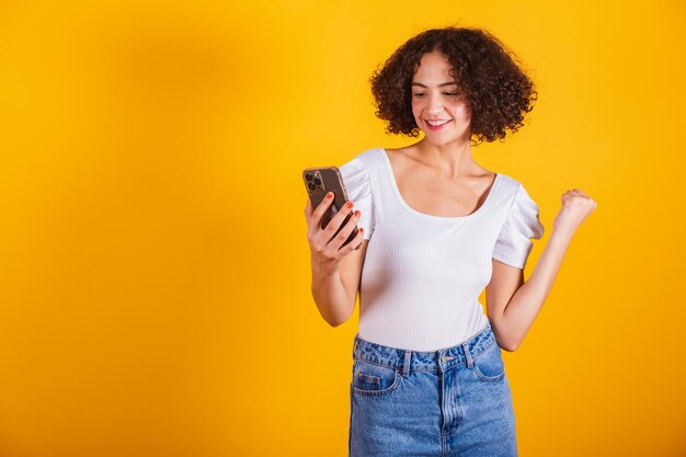 Modello caucasico in camicia bianca e jeans blu che tiene in mano uno smartphone per festeggiare