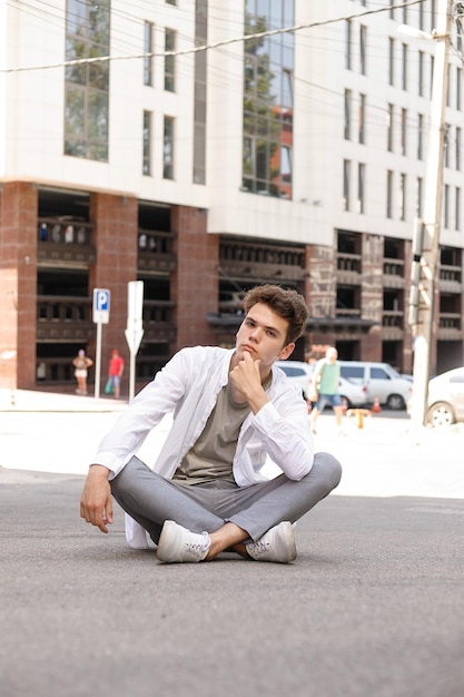 Modello del ragazzo con un taglio di capelli alla moda che posa all'aperto in una camicia bianca e pantaloni grigi. l'acconciatura alla moda riposa vicino a un moderno centro commerciale. Ragazzo attraente in strada in una giornata estiva.