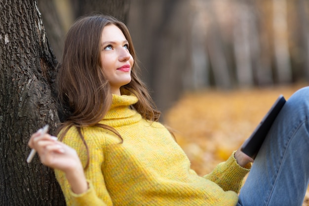 Modello della giovane donna nel parco di autunno con le foglie di acero gialle del fogliame. Moda per la stagione autunnale.