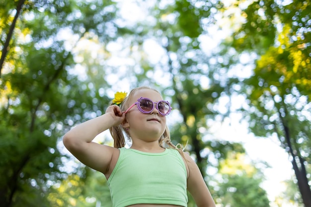 Modello di bambina in occhiali da sole sullo sfondo di alberi nel parco in posa per la fotocamera