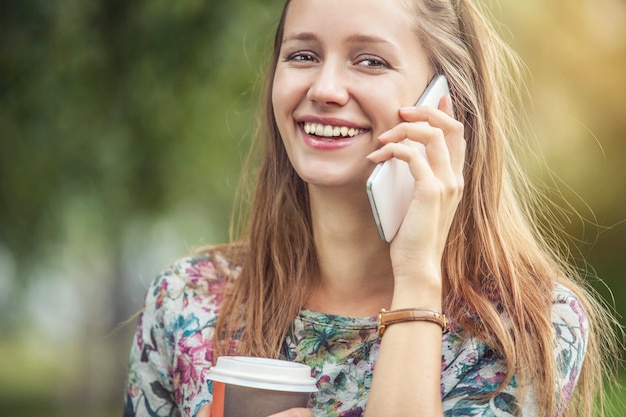 Modello di bella donna con caffè da asporto e un telefono nel parco. Stile, casual, bevanda, felicità, sole