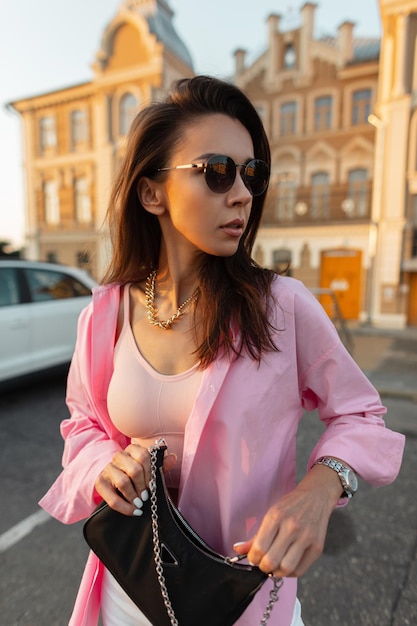 Modello di bella giovane donna alla moda con occhiali da sole alla moda in una camicia rosa alla moda e top che cammina in città e stringe la borsa