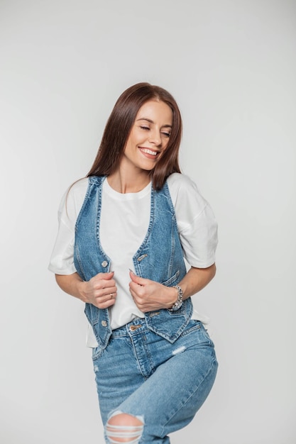 Modello di bella giovane donna allegra e carina con un sorriso carino in abiti di jeans di moda alla moda con una maglietta di gilet di jeans e jeans in posa su uno sfondo bianco in studio