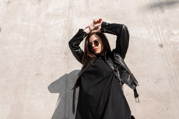 Modello di bella giovane ragazza caucasica cool con occhiali da sole hipster alla moda in eleganti vestiti neri con una giacca di pelle e felpa su uno sfondo di muro di cemento alla luce del sole