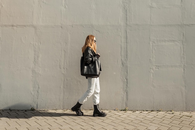 Modello di bella ragazza alla moda alla moda con occhiali da sole in abiti neri con giacca di pelle, jeans e stivali con una borsa che cammina vicino a un muro grigio in una soleggiata giornata primaverile