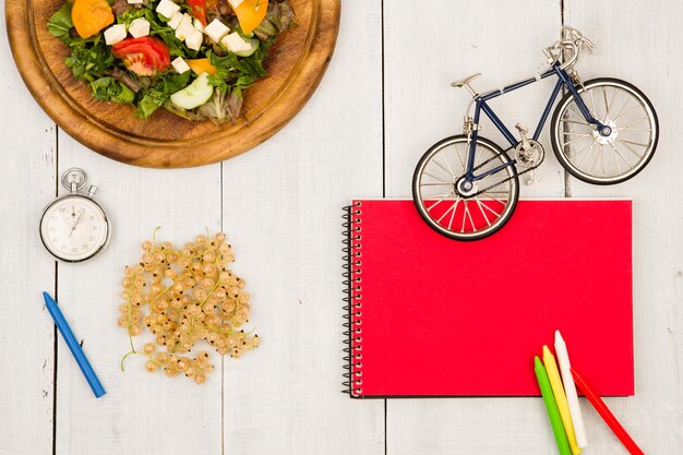 Modello di bicicletta insalata di verdure fresche rosso blocco note cronometro e ribes su un tavolo di legno bianco