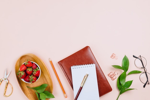 Modello di business flatlay con fragole su un vassoio di legno, quaderni, bicchieri, rami di ruscus e altri accessori su sfondo pastello con copyspace