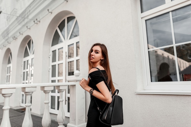 Modello di donna giovane e bella gioiosa in un elegante abito nero con uno zaino nero elegante in pelle viaggia per la città. Felice ragazza alla moda gode di una passeggiata su una strada. Moda giovanile di strada.