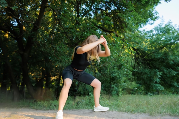 Modello di fitness ragazza snella in una tuta nera esegue squat tenendo le mani davanti a lei