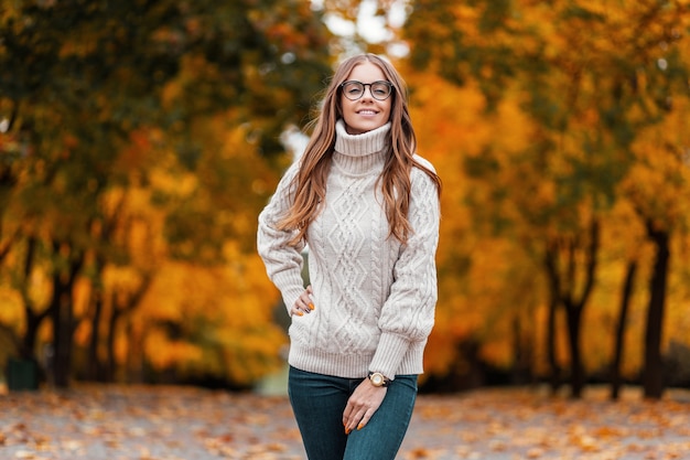 Modello di giovane donna felice in un maglione lavorato a maglia bianco in occhiali alla moda si trova nel parco e sorride positivamente sullo sfondo di alberi autunnali con fogliame arancione. Ragazza gioiosa hipster nella foresta.