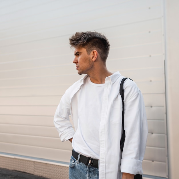 Modello di giovane uomo attraente in un'elegante camicia bianca in una maglietta in jeans blu alla moda con una borsa vintage si trova sulla strada