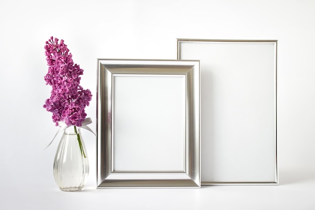 Modello di mockup con due cornici d'argento vuote e ramo di fiori lilla rosa in vaso di vetro su sfondo bianco.