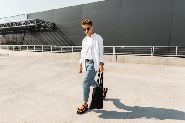 Modello di moda giovane americano in jeans alla moda in una camicia elegante in sandali alla moda in occhiali da sole con borsa cammina vicino a un edificio grigio.