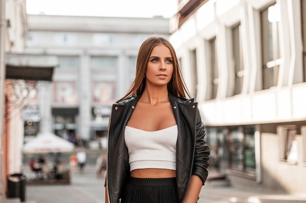 Modello di moda giovane donna attraente moderna in un'elegante camicia bianca in giacca di pelle nera alla moda gode di un sole splendente in una giornata autunnale all'aperto. La ragazza abbastanza urbana cammina sulla città.