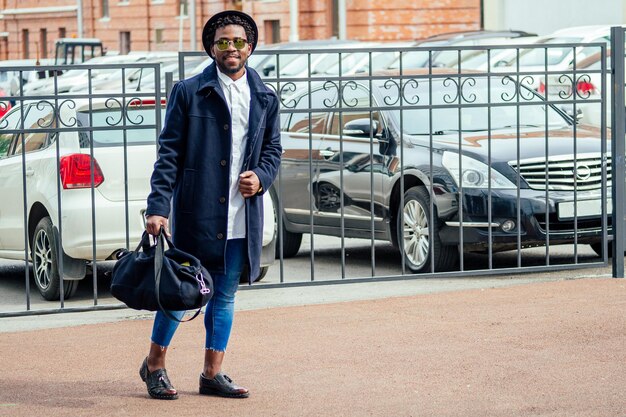 Modello di moda uomo afroamericano in cappotto e cappello jeans attillati che cammina lungo la strada della città vicino al parcheggio.