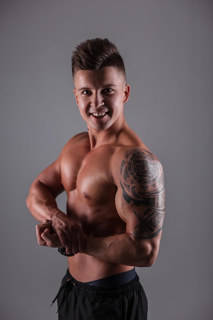 Modello di ragazzo atleta bodybuilder felice con corpo muscoloso nudo e tatuaggio che mostra i muscoli bicipiti su sfondo scuro