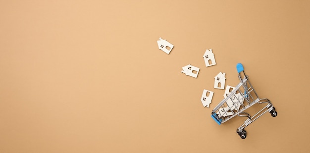 Modello di una casa in legno e di un carrello della spesa in miniatura in metallo su uno sfondo marrone chiaro, vista dall'alto. Concetto di ricerca casa per affitto, acquisto, mutuo