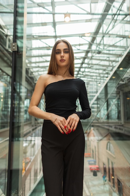 Modello elegante bella giovane donna in abiti eleganti neri con un top di moda e pantaloni vicino a una parete di vetro in un edificio commerciale