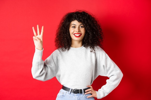 Modello femminile allegro che mostra il numero tre e sorride, fa un ordine, in piedi in felpa su sfondo rosso
