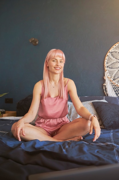 Modello femminile con lunghi capelli rosa in pigiama elegante si siede nella posa del loto sul letto comodo