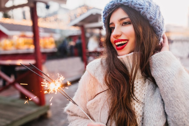 Modello femminile da sogno con capelli scuri e lucidi che si rilassano per strada con lo sparkler. Una bella donna indossa un cappello lavorato a maglia alla moda che celebra il nuovo anno con la luce del Bengala.