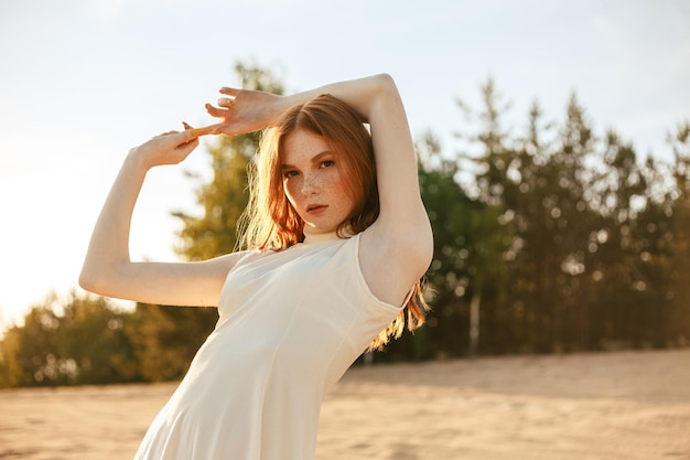 Modello femminile rosso sicuro in vestito bianco in piedi con le mani alzate in natura al tramonto