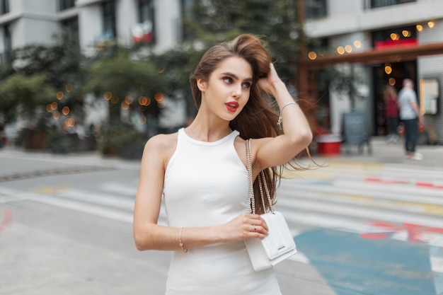 Modello glamour bella giovane ragazza elegante con le labbra rosse in un abito elegante alla moda con una borsa passeggiate in città raddrizza i capelli