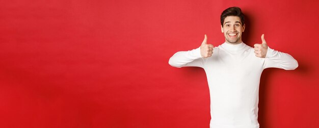 Modello maschio allegro di bell'aspetto in maglione bianco che mostra il pollice in su in segno di approvazione come qualcosa di buono...