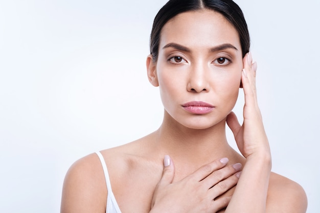 Modello professionale. Bella giovane donna dai capelli scuri che tiene sulle sue mani vicino al viso mentre si tiene l'altra mano sul petto e posa contro un muro bianco