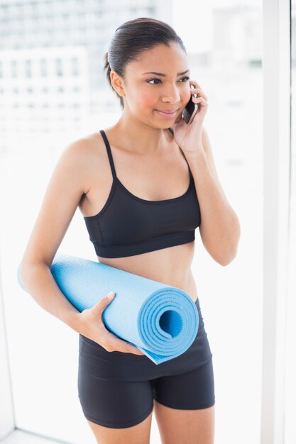 Modello sano dai capelli scuri in abiti sportivi che trasportano un materassino e fare una telefonata