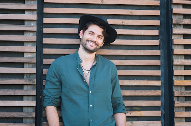 Modello uomo sorridente vestito con cappello nero e camicia verde, foto all'aperto contro la parete di legno