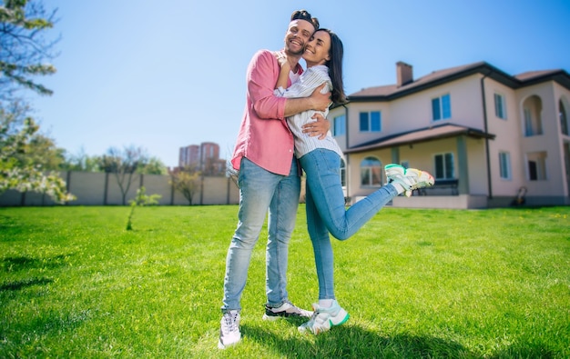 Moderna bella giovane coppia eccitata innamorata in piedi di fronte alla nuova grande casa con le chiavi in mano e abbracciata mentre celebrano questo acquisto