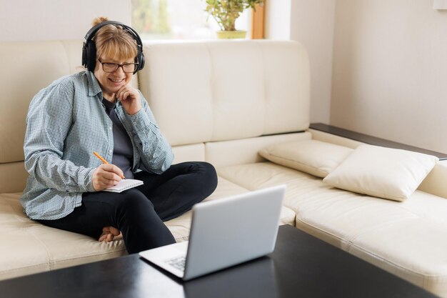 Moderna donna bionda di mezza età in cuffia segui un corso online o un corso di formazione sul computer a casa Una donna caucasica matura intelligente in auricolari guarda il webinar prendi nota di uno studio distante sul laptop