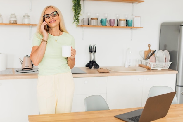 Moderna donna di mezza età attraente che parla al telefono e beve caffè in cucina a casa