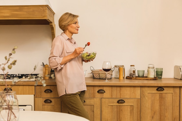 Moderna donna matura invecchiata senior che mangia insalata verde fresca e verdure in cucina, sorridendo felice. Concetto di stile di vita sano.