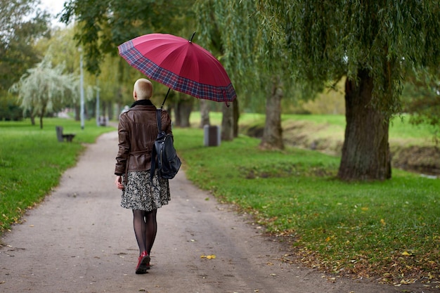 Moderna ed elegante donna bionda dai capelli corti cammina nel parco autunnale con ombrello rosso Vista dal retro