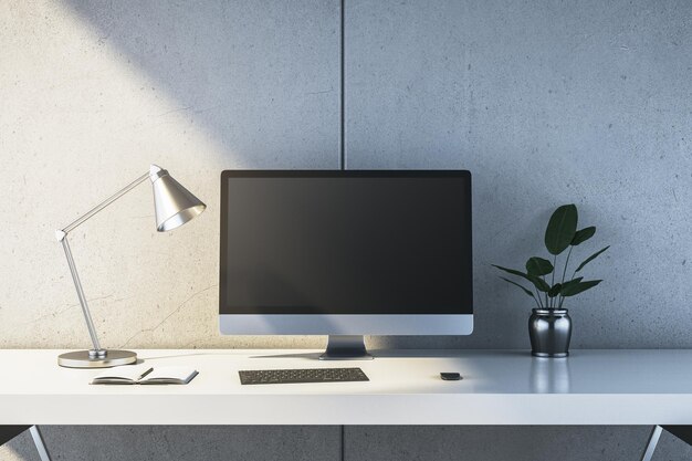 Moderna scrivania bianca con computer desktop, lampada, quaderno e pianta contro un muro di cemento grigio minimalista in un interno luminoso Rendering 3D