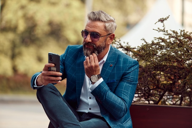 Moderno uomo d'affari senior che utilizza lo smartphone per riunioni online mentre è seduto sulla panchina.